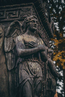 A detailed angelic sculpture stands against a background of intricate architectural designs, with weathered textures and a somber expression. The figure has wings and its arms are crossed over its chest. The surroundings include hints of foliage with autumn colors, adding depth and contrast to the stonework.