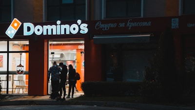 A nighttime street view of a Domino's pizza store. The store is brightly lit, with the illuminated Domino's sign prominently displayed. There are people entering or exiting the store, and an 'OPEN' sign is visible in the window. The surrounding area is darker, with minimal lighting on the street and bushes nearby.