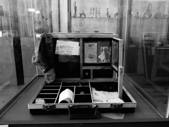 An open briefcase is displayed on a table inside a glass case. The internal compartments of the briefcase are partly filled with various items, including documents, a printed receipt or label, and other small items. Behind the glass case, shelves exhibit pottery or artifact pieces, faintly visible in the background.