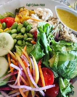 A colorful and vibrant salad composed of various fresh ingredients including leafy greens, sliced cucumbers, cherry tomatoes, edamame, shredded carrots, and thinly sliced radishes. There are also roasted vegetables such as cauliflower and squash, served with a side of quinoa and a small bowl of yellow dressing. The dish is presented in a white bowl labeled Café L'Etoile 1903.