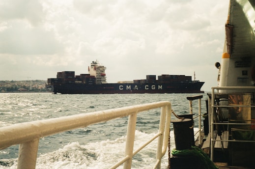 A large cargo ship with the letters 'CMA CGM' on its side is sailing on the ocean. The scene includes part of another boat in the foreground, with ropes and railings visible. The sky is cloudy, and land is visible in the distance.