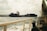 A large cargo ship with the letters 'CMA CGM' on its side is sailing on the ocean. The scene includes part of another boat in the foreground, with ropes and railings visible. The sky is cloudy, and land is visible in the distance.