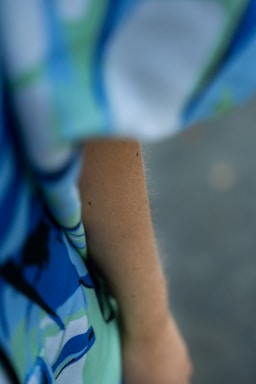 A close-up view of a person's arm wearing a garment with a colorful, abstract pattern. The fabric features shades of blue, green, and hints of white and black, creating a vibrant, flowing design. The focus is on the arm and the fabric, with a soft, blurred background.