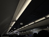 The interior of an airplane cabin with overhead storage compartments and illuminated lights. Passengers are seated below the compartments in a dimly lit setting.