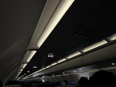 The interior of an airplane cabin with overhead storage compartments and illuminated lights. Passengers are seated below the compartments in a dimly lit setting.