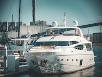 A luxurious white yacht docked at a marina, surrounded by industrial buildings and structures in the background. The yacht is equipped with various modern equipment and is stationed in calm waters.