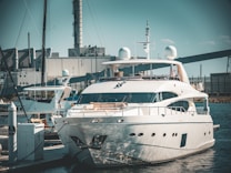 A luxurious white yacht docked at a marina, surrounded by industrial buildings and structures in the background. The yacht is equipped with various modern equipment and is stationed in calm waters.
