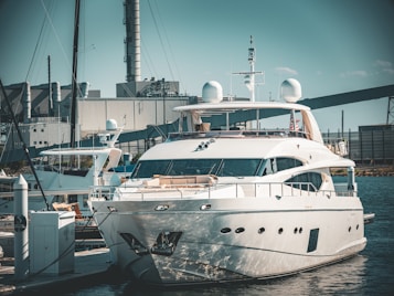 A luxurious white yacht docked at a marina, surrounded by industrial buildings and structures in the background. The yacht is equipped with various modern equipment and is stationed in calm waters.