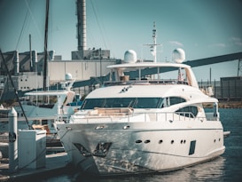 A luxurious white yacht docked at a marina, surrounded by industrial buildings and structures in the background. The yacht is equipped with various modern equipment and is stationed in calm waters.