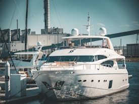 A luxurious white yacht docked at a marina, surrounded by industrial buildings and structures in the background. The yacht is equipped with various modern equipment and is stationed in calm waters.