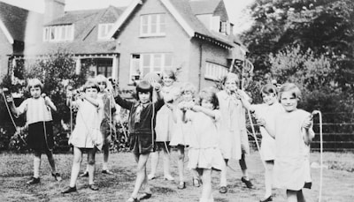 A group of children are playing outside with skipping ropes on a grassy area. The background features a large brick house with multiple windows and a garden with bushes and trees.