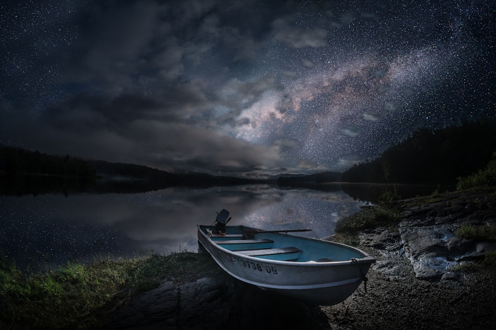 a boat sitting on the shore of a lake under a night sky filled with stars