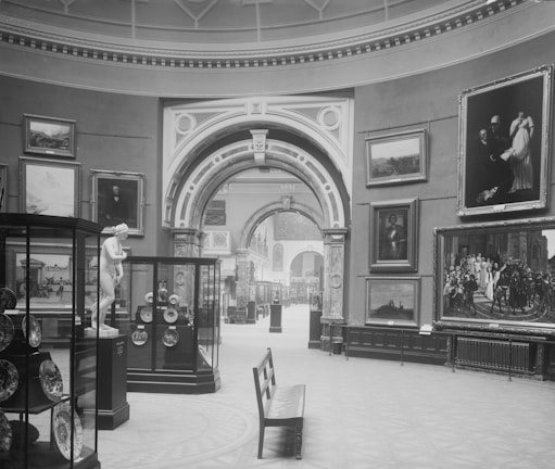 A black and white photograph of an art gallery interior displaying various paintings and sculptures. The room features high ceilings with ornate architectural details, including arches and columns. Several framed artworks are hung on the walls, and glass display cases contain plates and artifacts. A bench is positioned prominently in the center of the room.