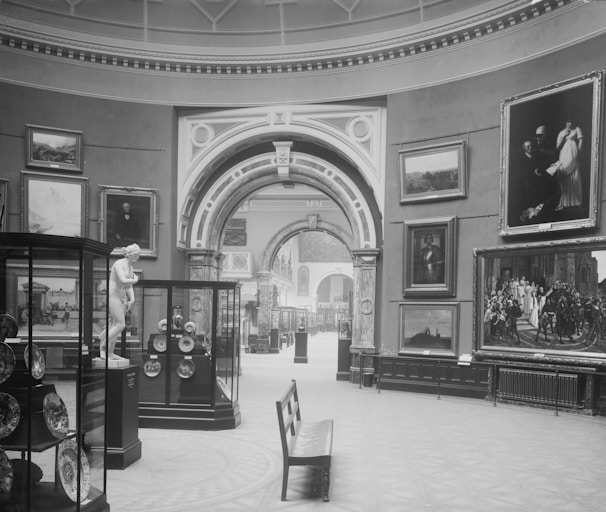 A black and white photograph of an art gallery interior displaying various paintings and sculptures. The room features high ceilings with ornate architectural details, including arches and columns. Several framed artworks are hung on the walls, and glass display cases contain plates and artifacts. A bench is positioned prominently in the center of the room.