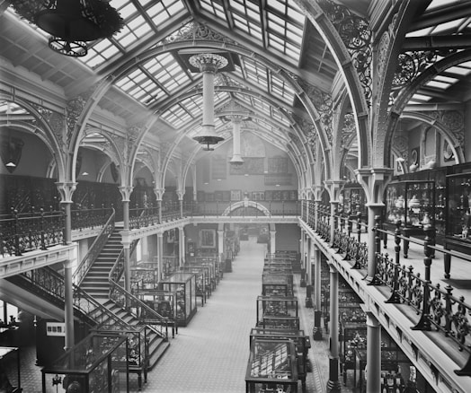 A grand, ornate interior of a historical building with high arched ceilings and intricate ironwork. The space is filled with glass display cases containing various artifacts. A large staircase on the left side connects two levels, while natural light streams in through the skylights, illuminating the detailed architectural features.