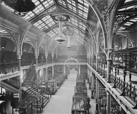 A grand, ornate interior of a historical building with high arched ceilings and intricate ironwork. The space is filled with glass display cases containing various artifacts. A large staircase on the left side connects two levels, while natural light streams in through the skylights, illuminating the detailed architectural features.