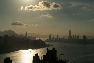 A city skyline in silhouette with a large body of water reflecting sunlight. Tall buildings, including a distinctive tower, are set against a mountain backdrop and a sunlit sky with scattered clouds.