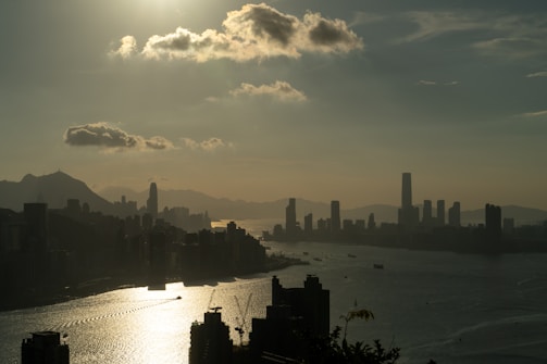 A city skyline in silhouette with a large body of water reflecting sunlight. Tall buildings, including a distinctive tower, are set against a mountain backdrop and a sunlit sky with scattered clouds.