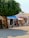 A street scene featuring a quaint shop with various artworks and crafts displayed outside. A large tree offers shade, and colorful clothing hangs on racks. The shop appears to be small and charming, with a rustic feel, enhanced by potted plants and an inviting entrance.