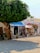 A street scene featuring a quaint shop with various artworks and crafts displayed outside. A large tree offers shade, and colorful clothing hangs on racks. The shop appears to be small and charming, with a rustic feel, enhanced by potted plants and an inviting entrance.