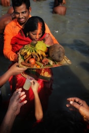 A woman dressed in traditional red attire stands in water holding a large basket filled with fruits and vegetables, including bananas, coconuts, and other offerings. A man behind her supports her, and several hands are visible reaching towards the offerings. The scene appears to be a ritual or religious ceremony.