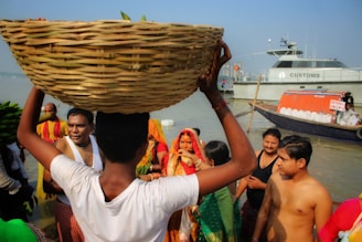 A group of people, some dressed in colorful attire, are standing near a river. One person is balancing a large woven basket on their head. In the background, a boat with a 'CUSTOMS' sign is docked on the water. The scene appears lively, possibly indicating a cultural or religious gathering.