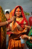 A woman dressed in a traditional orange and red sari with intricate patterns stands near a body of water. She has a decorative nose ring and red vermilion applied on her forehead. Multiple hands are seen, one applying sindoor and others holding various items. The background includes more people and greenery.