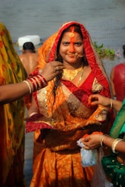 A woman dressed in a traditional orange and red sari with intricate patterns stands near a body of water. She has a decorative nose ring and red vermilion applied on her forehead. Multiple hands are seen, one applying sindoor and others holding various items. The background includes more people and greenery.