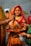 A woman dressed in a traditional orange and red sari with intricate patterns stands near a body of water. She has a decorative nose ring and red vermilion applied on her forehead. Multiple hands are seen, one applying sindoor and others holding various items. The background includes more people and greenery.