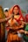 A woman dressed in a traditional orange and red sari with intricate patterns stands near a body of water. She has a decorative nose ring and red vermilion applied on her forehead. Multiple hands are seen, one applying sindoor and others holding various items. The background includes more people and greenery.