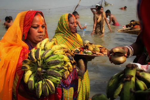Several women are dressed in colorful traditional sarees, holding offerings such as bananas and plates of fruit and other items. They are standing in a body of water, likely performing a ritual. In the background, more people are partially immersed in water, suggesting a communal or religious practice, possibly a festival.