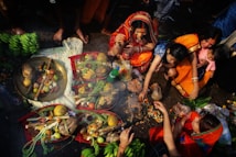 A group of people are engaged in a cultural or religious ritual. They are gathered around several baskets filled with fruits and vegetables, as well as a brass plate containing offerings. The women are wearing colorful traditional attire, and some are making gestures associated with the ritual.