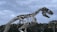 A large metal dinosaur skeleton sculpture is prominently displayed against a backdrop of cloudy skies and trees. The impressive and detailed structure is reminiscent of a prehistoric creature, showcasing sharp teeth and elongated limbs. The setting appears to be an outdoor area with a few buildings visible in the distance.
