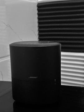 A black Bose speaker is placed on a reflective surface. The background consists of a wall with a combination of a marble pattern and black acoustic foam panels.