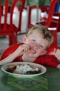A young child is smiling and enjoying a meal of rice and seafood. The background features red chairs, and the table is covered with a green tablecloth.