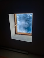 A rectangular skylight window set in a dark ceiling shows raindrops on the glass with a cloudy blue sky visible outside.