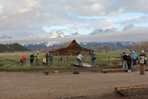 A group of people with cameras and tripods set up near a rustic wooden barn on a grassy field. In the background, snow-capped mountains rise under a partly cloudy sky. The setting suggests a tranquil, scenic landscape ideal for photography.