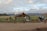 A group of people with cameras and tripods set up near a rustic wooden barn on a grassy field. In the background, snow-capped mountains rise under a partly cloudy sky. The setting suggests a tranquil, scenic landscape ideal for photography.