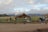 A group of people with cameras and tripods set up near a rustic wooden barn on a grassy field. In the background, snow-capped mountains rise under a partly cloudy sky. The setting suggests a tranquil, scenic landscape ideal for photography.