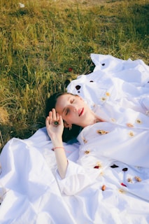 A person lies peacefully on a bed of white fabric adorned with scattered flowers. The surrounding terrain is grassy and slightly dry, indicating a serene outdoor setting. The individual seems to be in a state of relaxation or contemplation.