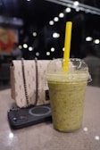A refreshing-looking green smoothie in a plastic cup with a yellow straw is placed on a table. Next to it are a stylish beige purse with a chain and a black smartphone. The background is a dimly lit indoor setting with blurred lights and a few people.