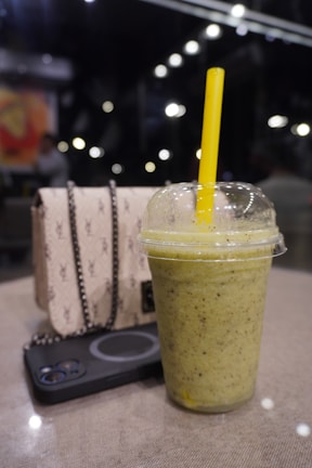 A refreshing-looking green smoothie in a plastic cup with a yellow straw is placed on a table. Next to it are a stylish beige purse with a chain and a black smartphone. The background is a dimly lit indoor setting with blurred lights and a few people.