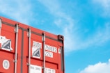 A red shipping container with various labels is positioned against a bright blue sky with scattered clouds. The labels include corrosion warnings and numerical identifiers. The container's metal rivets and structures are visible, contributing to an industrial aesthetic.