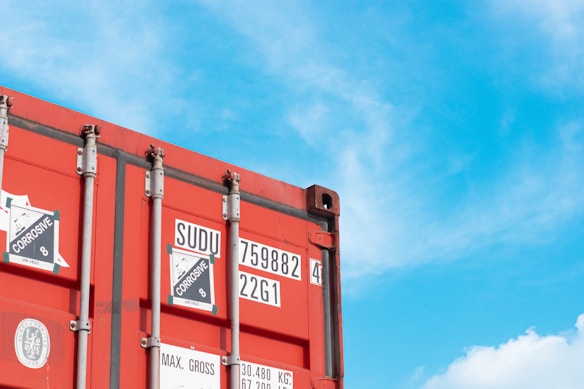 A red shipping container with various labels is positioned against a bright blue sky with scattered clouds. The labels include corrosion warnings and numerical identifiers. The container's metal rivets and structures are visible, contributing to an industrial aesthetic.