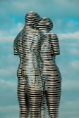 Two interlocking metallic sculptures resemble human figures, with a backdrop of a clear sky. The sculptures consist of horizontal metal layers stacked to form seamless, intertwined silhouettes. The figures appear to be embracing, conveying a sense of connection and unity.