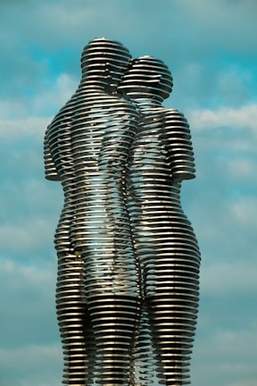 Two interlocking metallic sculptures resemble human figures, with a backdrop of a clear sky. The sculptures consist of horizontal metal layers stacked to form seamless, intertwined silhouettes. The figures appear to be embracing, conveying a sense of connection and unity.