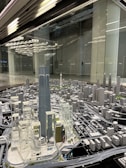 A detailed architectural model of a city, featuring numerous high-rise buildings, roads, and infrastructure. The centerpiece is a tall, prominent skyscraper surrounded by other modern structures. The setting seems to be a showroom or exhibition area with reflective surfaces and ceiling lights.