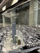 A detailed architectural model of a city, featuring numerous high-rise buildings, roads, and infrastructure. The centerpiece is a tall, prominent skyscraper surrounded by other modern structures. The setting seems to be a showroom or exhibition area with reflective surfaces and ceiling lights.