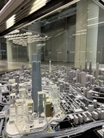 A detailed architectural model of a city, featuring numerous high-rise buildings, roads, and infrastructure. The centerpiece is a tall, prominent skyscraper surrounded by other modern structures. The setting seems to be a showroom or exhibition area with reflective surfaces and ceiling lights.