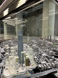 A detailed architectural model of a city, featuring numerous high-rise buildings, roads, and infrastructure. The centerpiece is a tall, prominent skyscraper surrounded by other modern structures. The setting seems to be a showroom or exhibition area with reflective surfaces and ceiling lights.